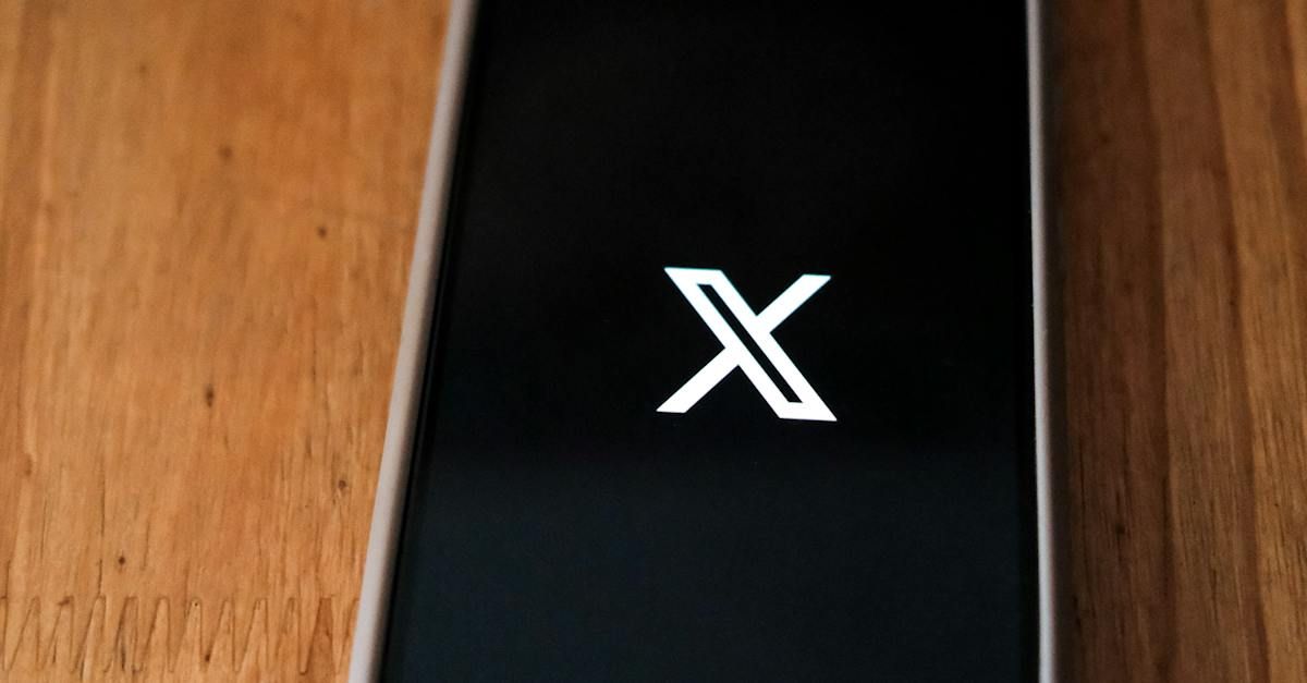 A cell phone with the letter x on the screen is sitting on a wooden table.