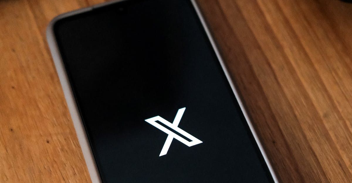 A cell phone with the letter x on the screen is sitting on a wooden table.