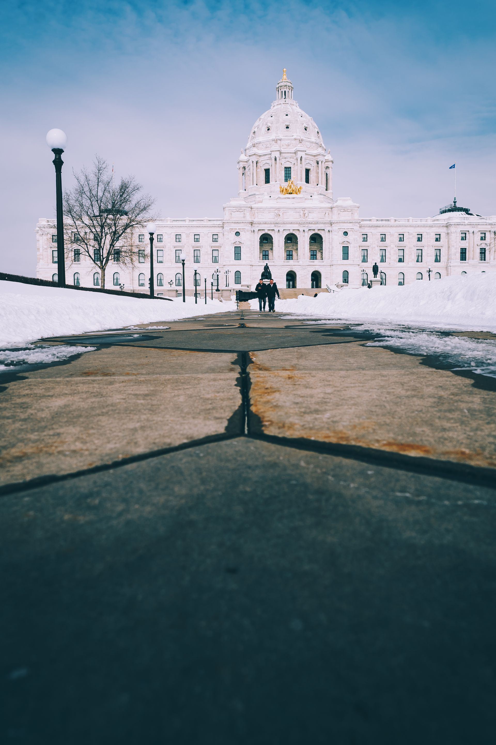 A person walks towards the white Minnesota State Capitol building in winter, snow on the ground. Blue sky overhead.