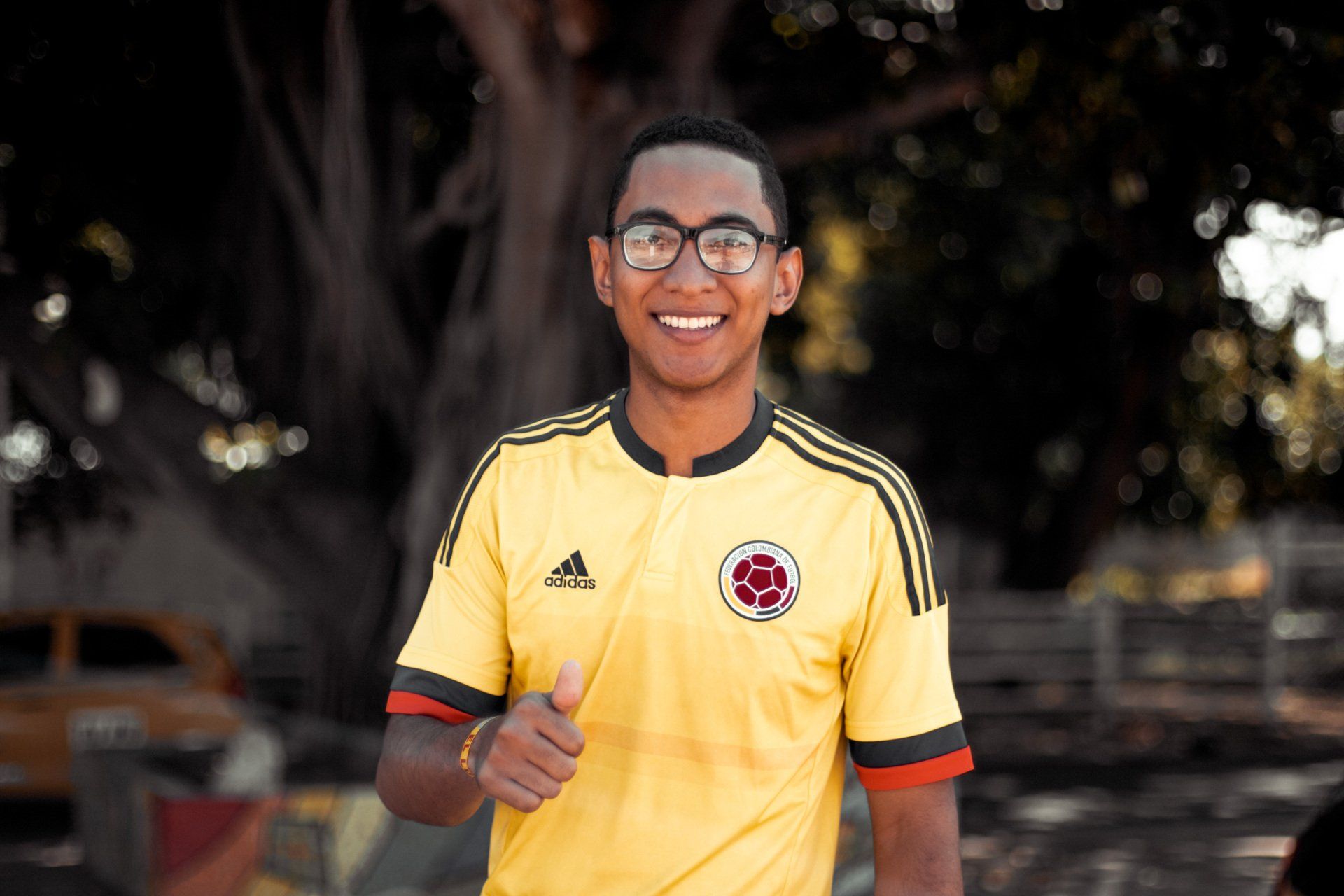 Man in yellow Colombia jersey with glasses smiles and gives a thumbs up outdoors.