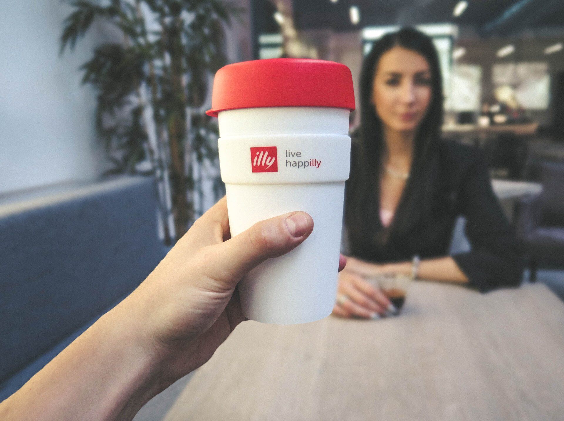 a person is holding a cup of illy coffee in front of a woman sitting at a table .
