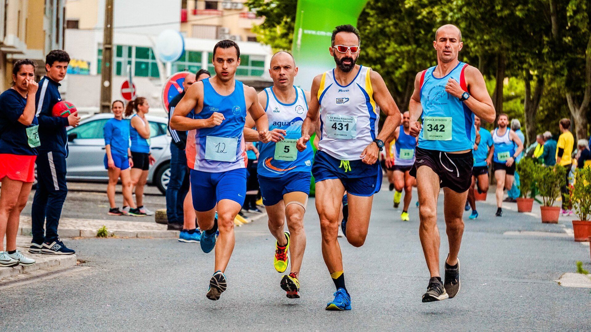 Runners in blue, white, and yellow shirts sprinting on a street. Spectators watch in the background.