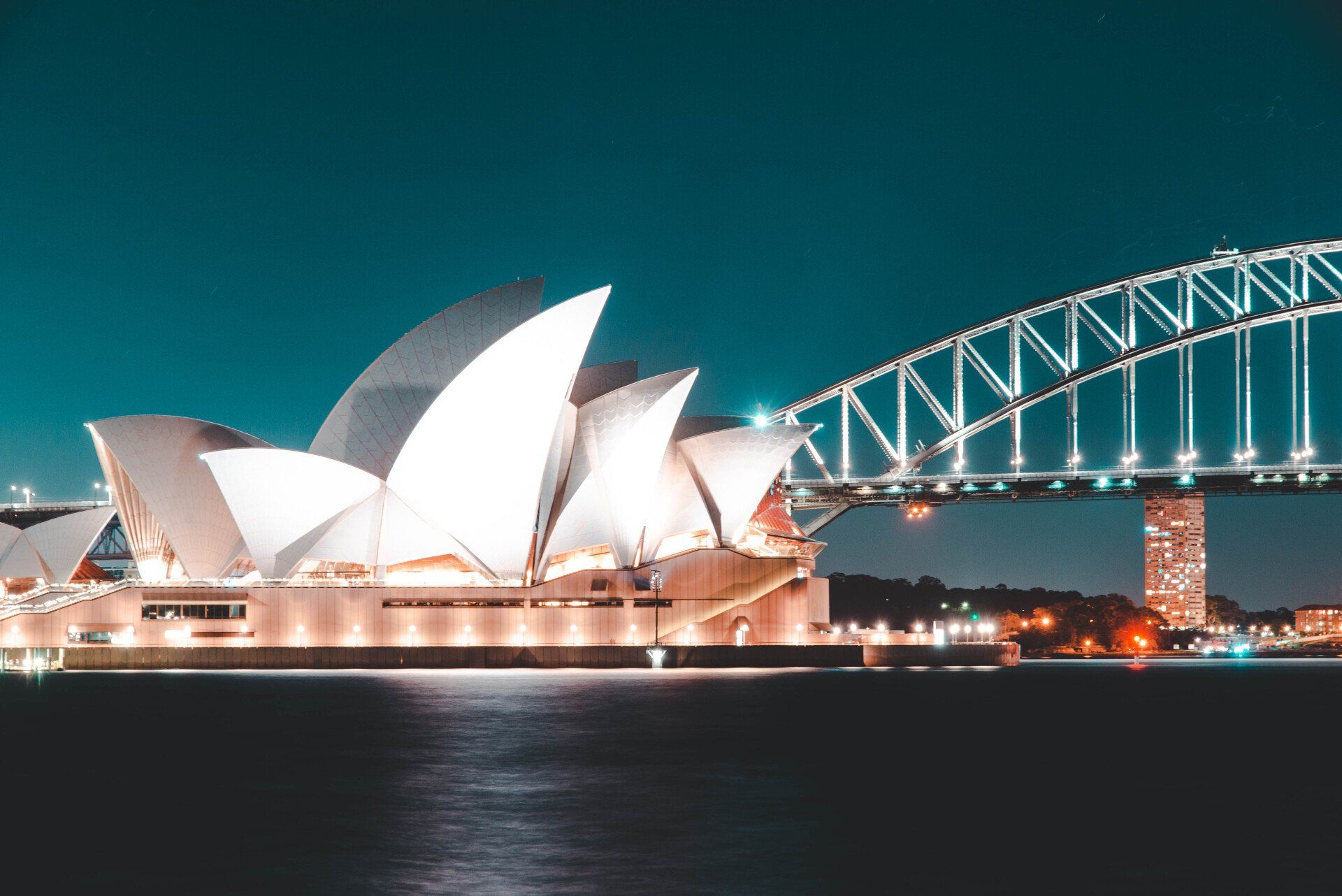 The sydney opera house is lit up at night with a bridge in the background.