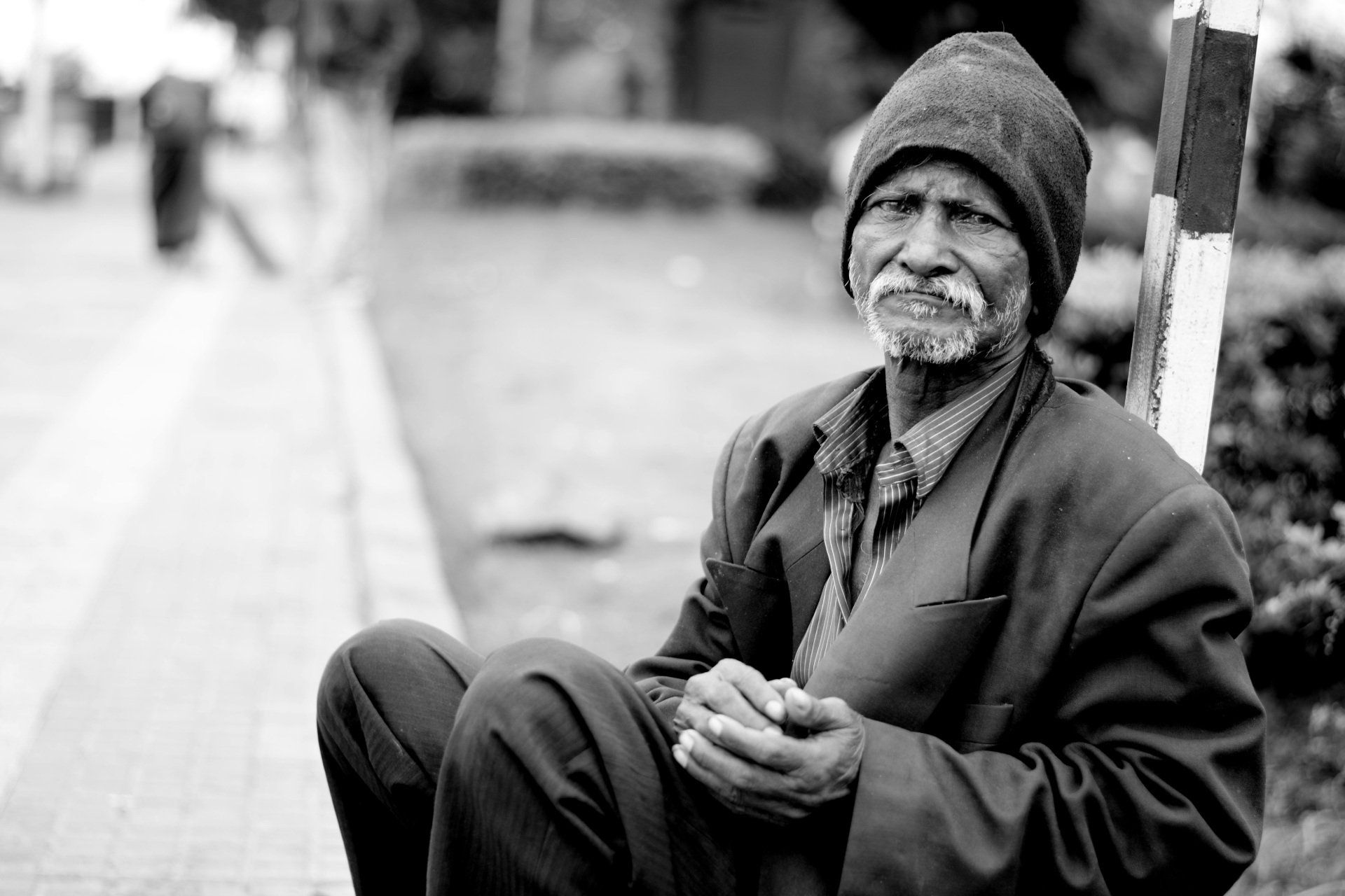A black and white photo of an elderly man sitting on the sidewalk.