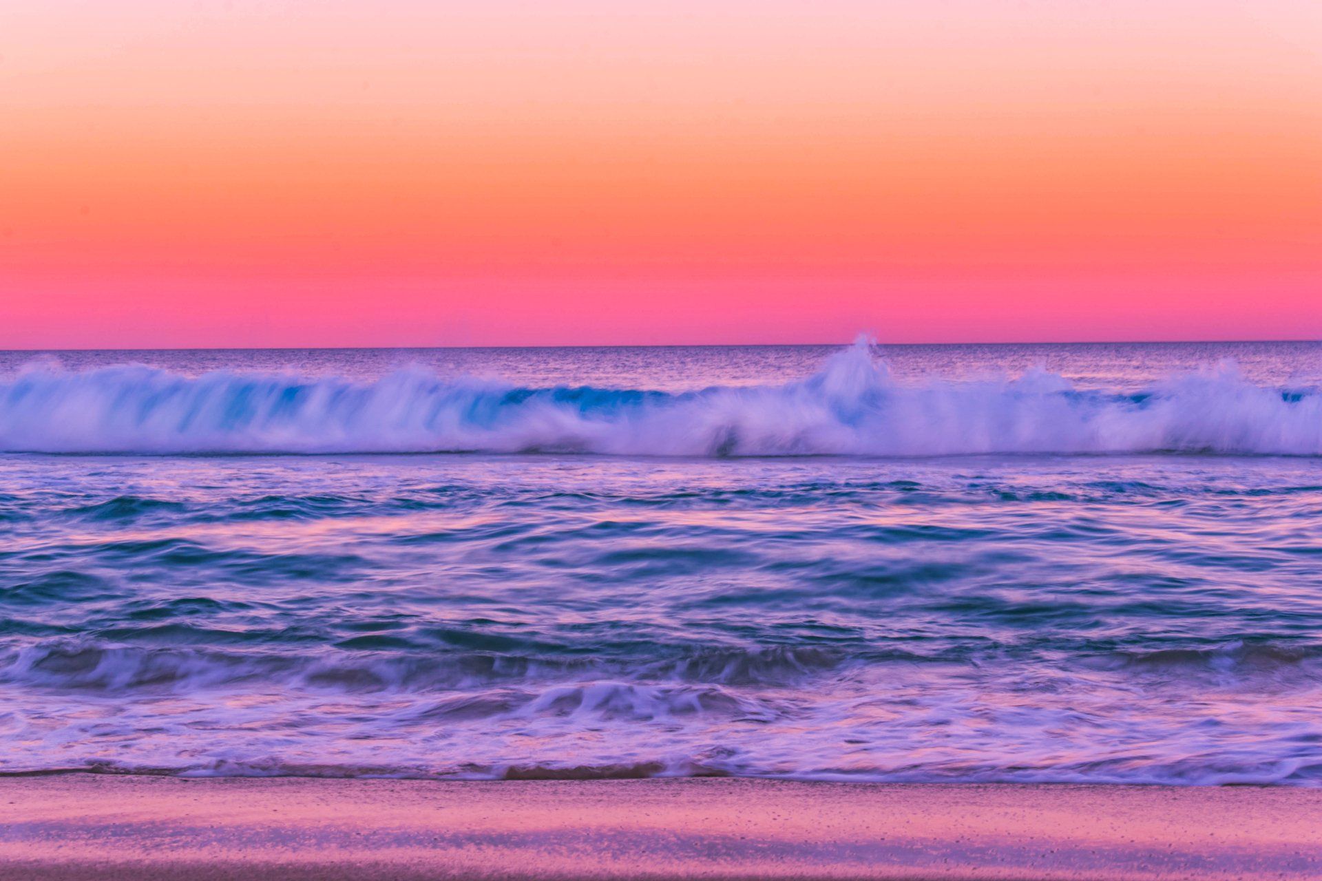 A pink and purple sunset over the ocean with waves crashing on the beach.