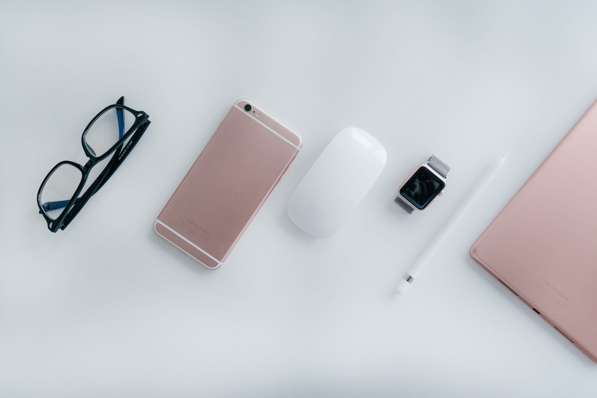 Black glasses, pink phone, white mouse, smart watch, pen, and laptop on white.