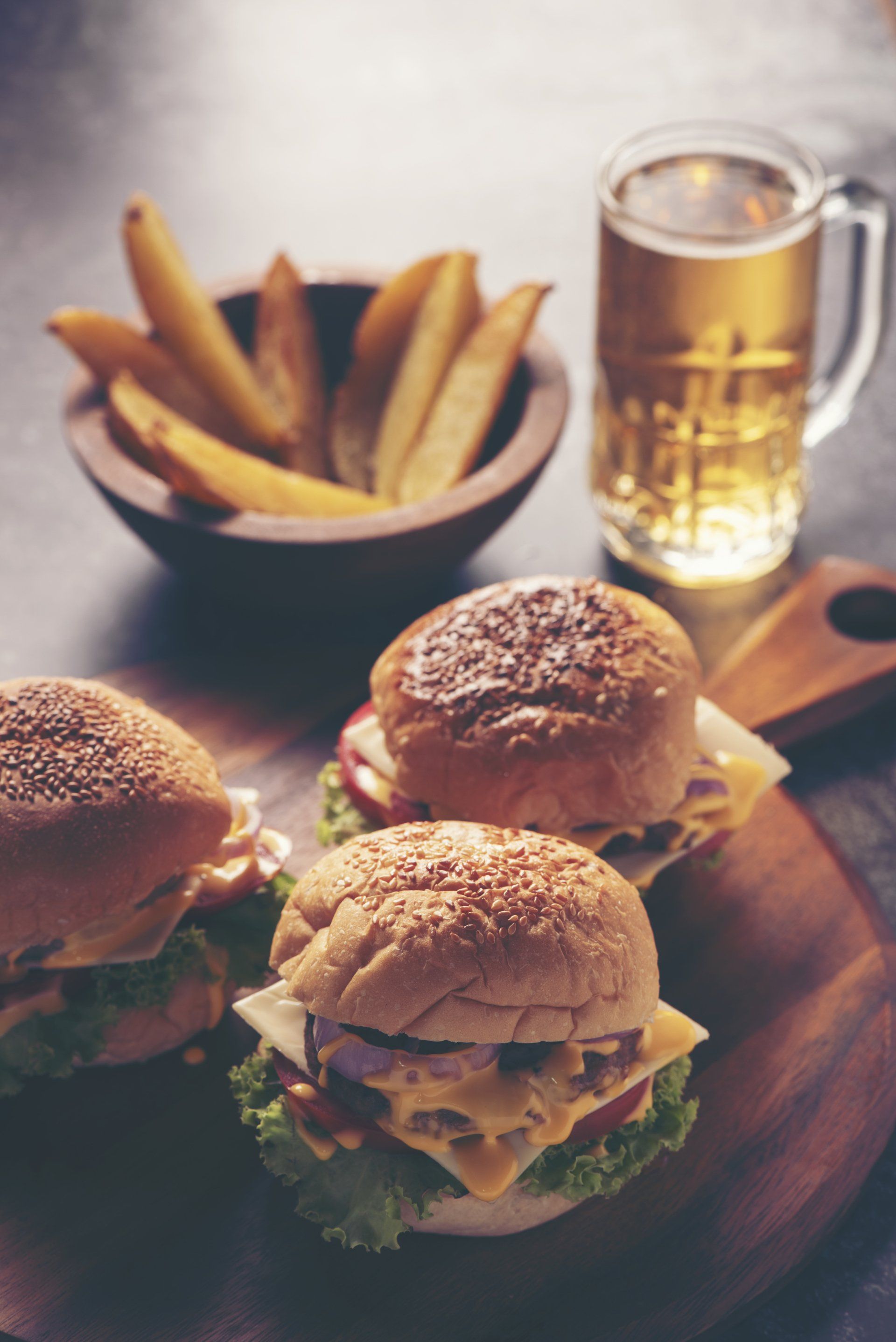 Three hamburgers are sitting on a wooden cutting board next to a bowl of french fries and a glass of beer.