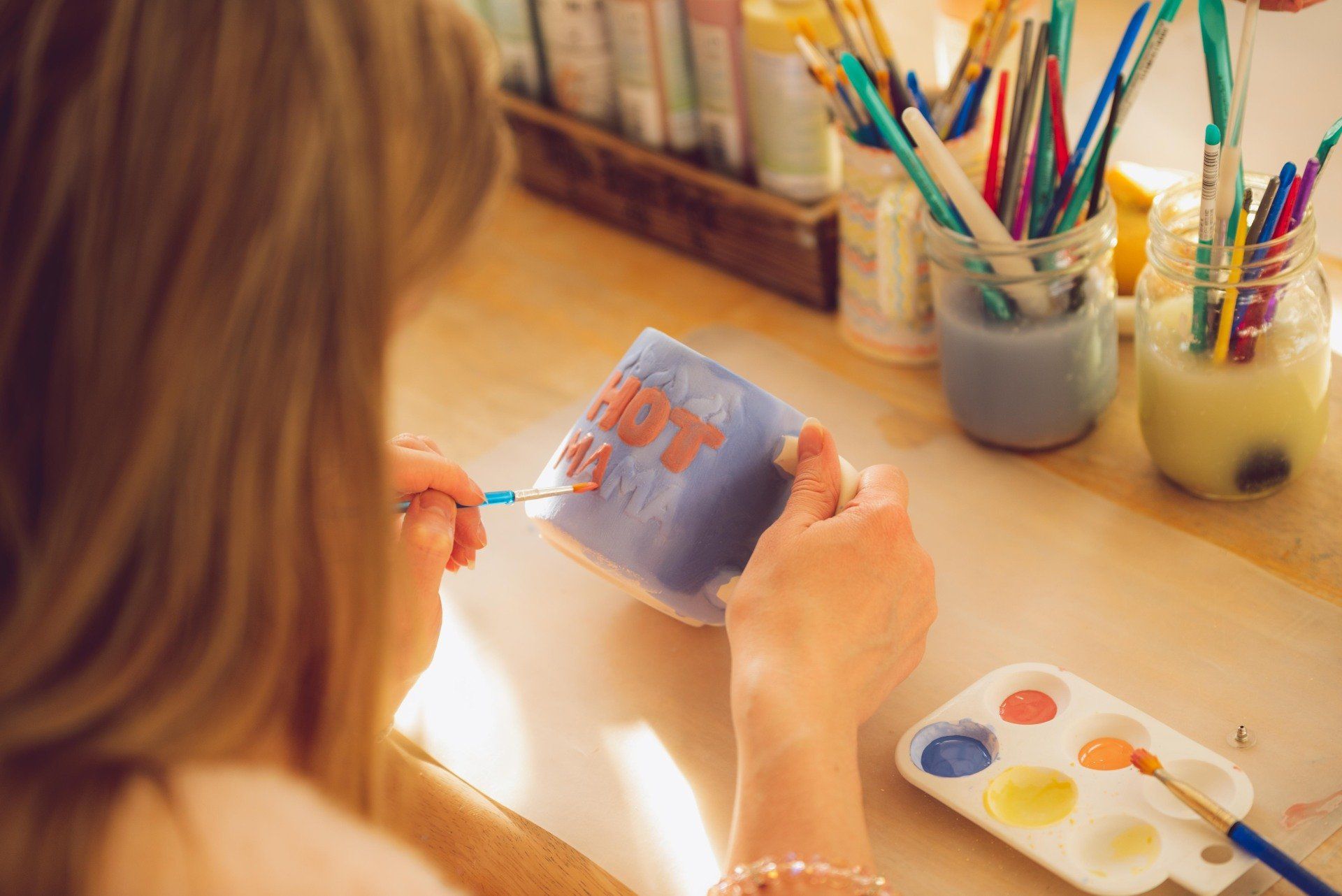 A woman is painting a mug with a brush on a table.