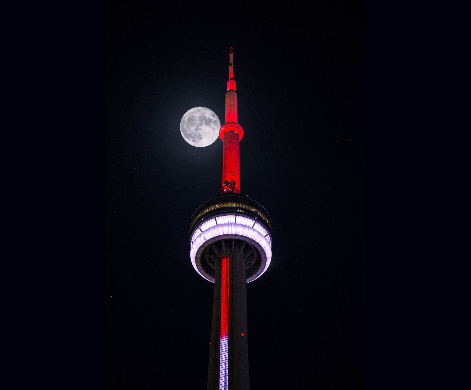 The cn tower is lit up at night with a full moon in the background.