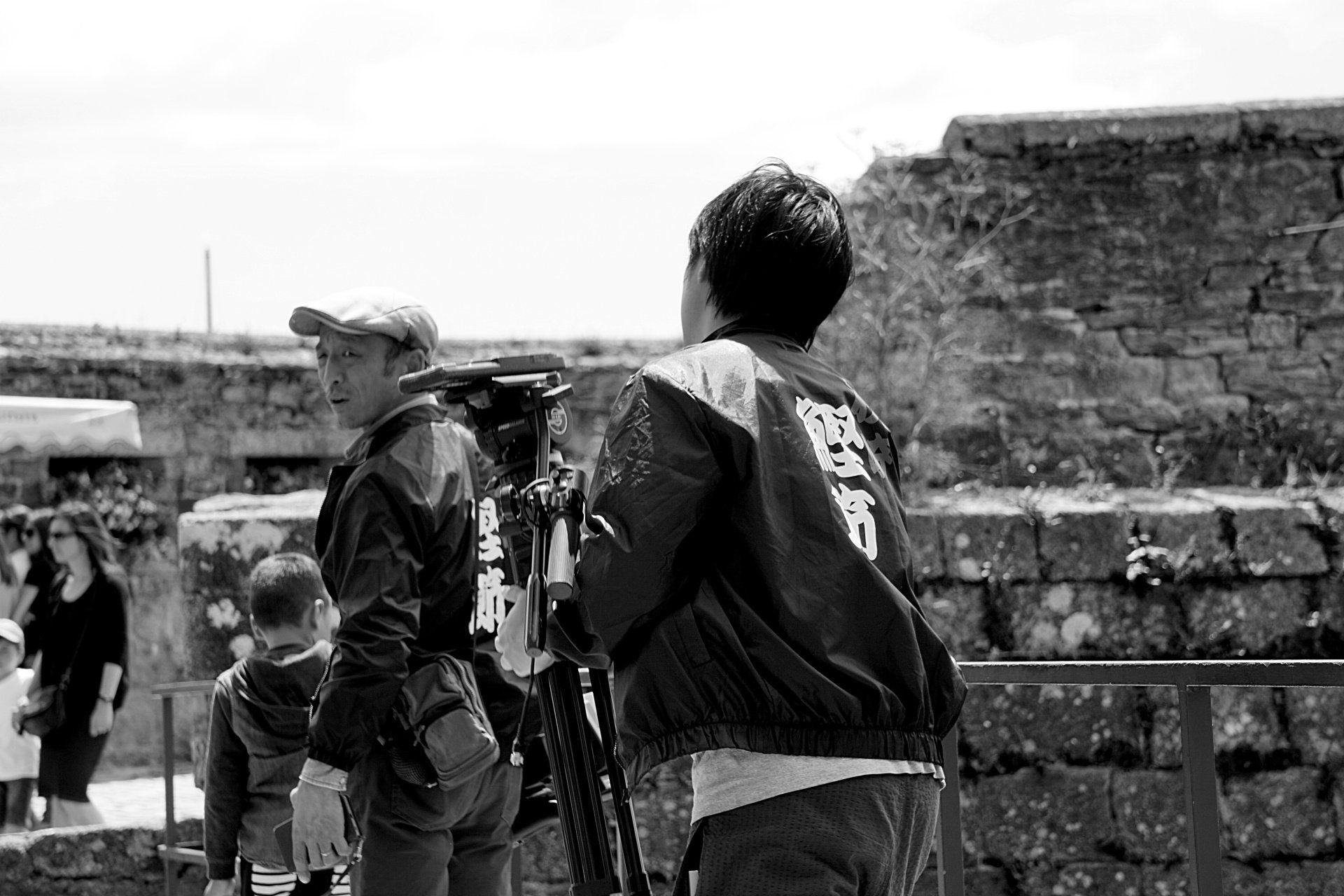 A man is holding a camera on a tripod in front of a stone wall.