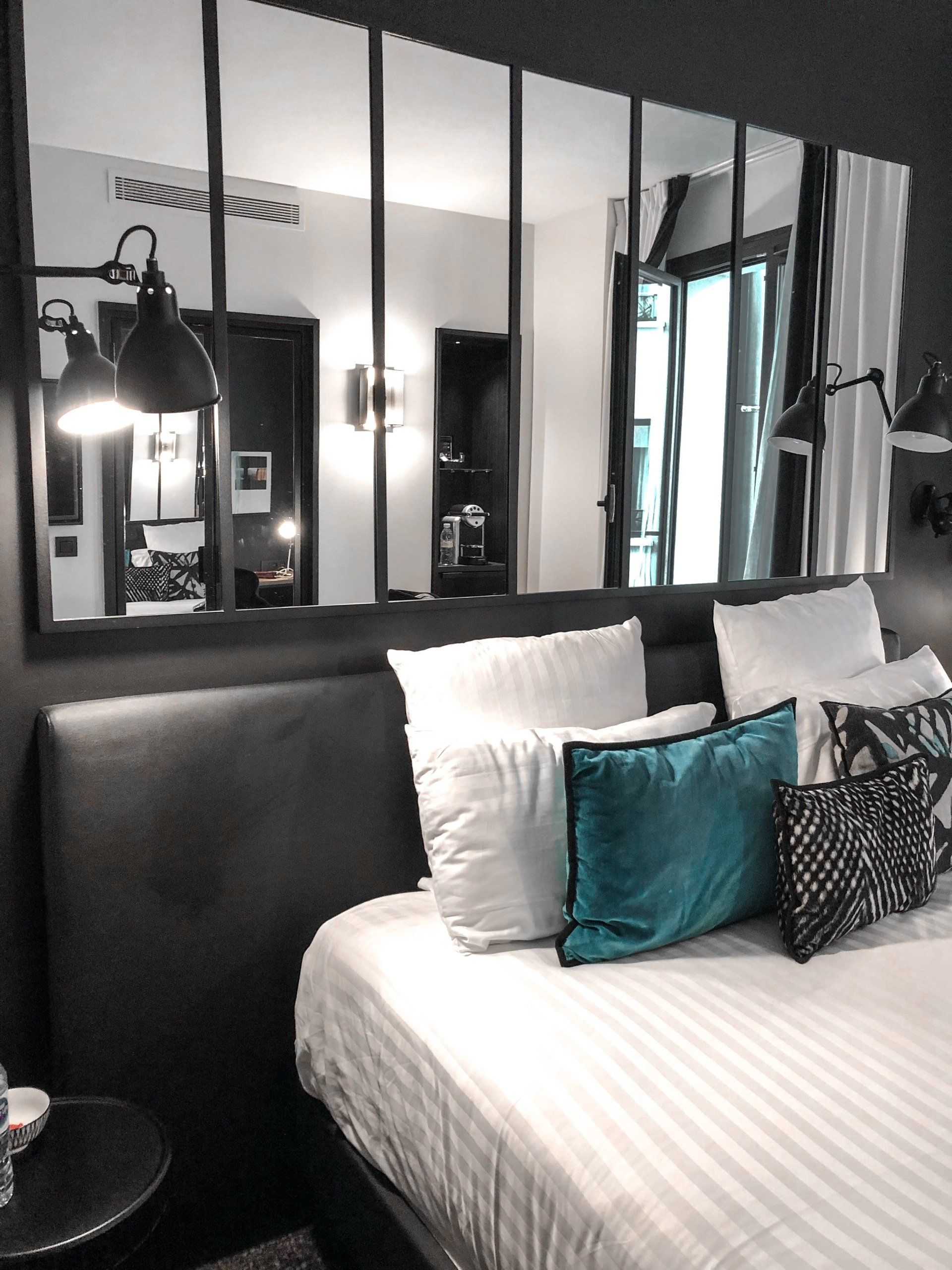 Bedroom with bed, large black-framed mirror, white bedding, teal and patterned pillows, black lamps, and dark gray walls.