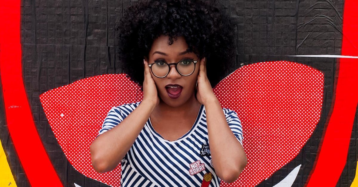 A woman wearing glasses is covering her ears in front of a graffiti wall.