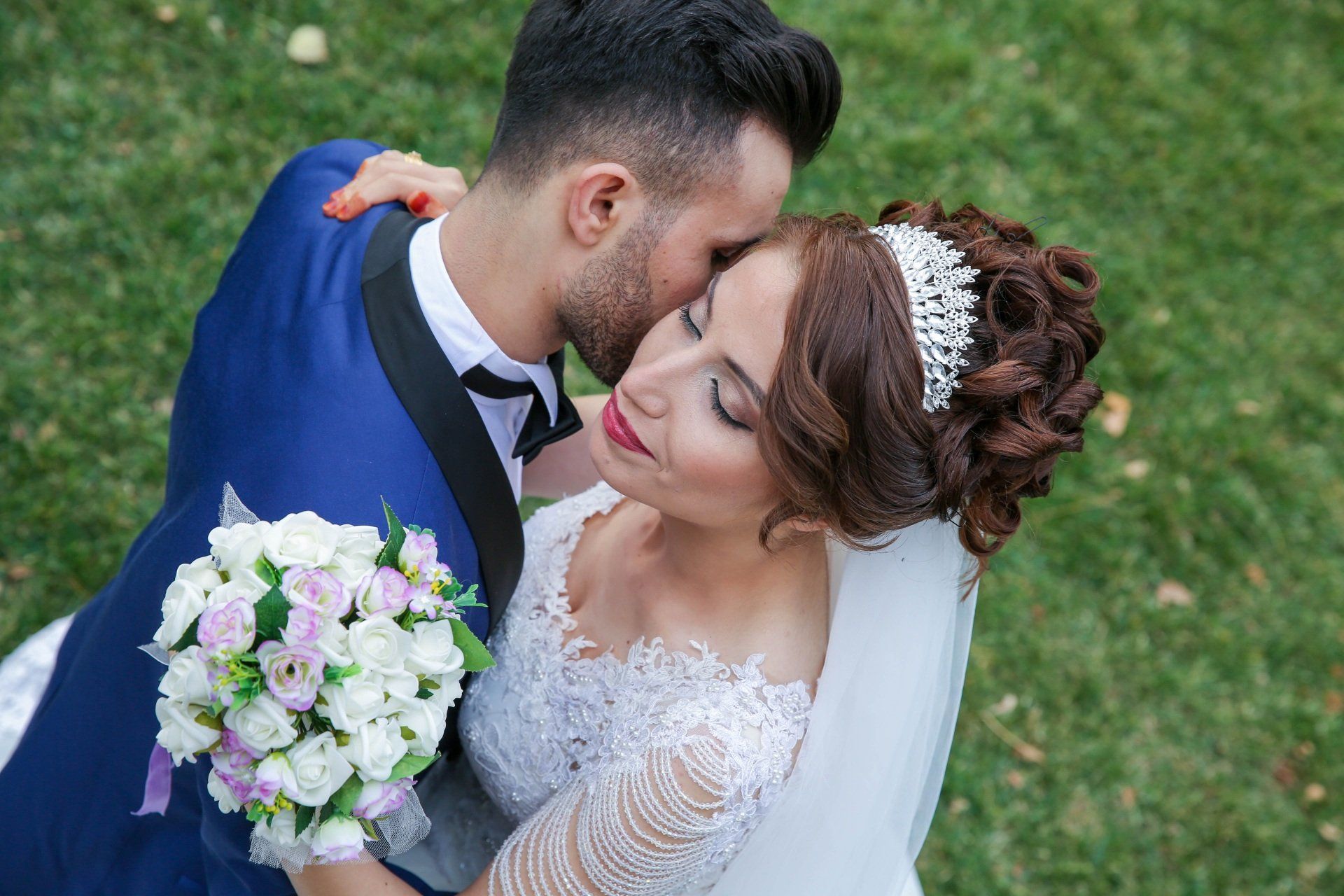 A bride and groom are kissing in the grass on their wedding day.