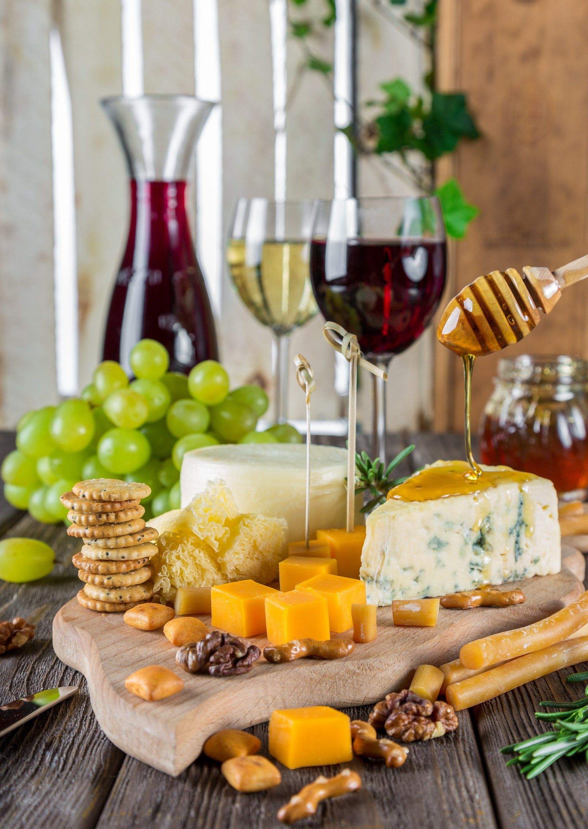 a wooden cutting board topped with cheese , crackers , honey , grapes and wine glasses .