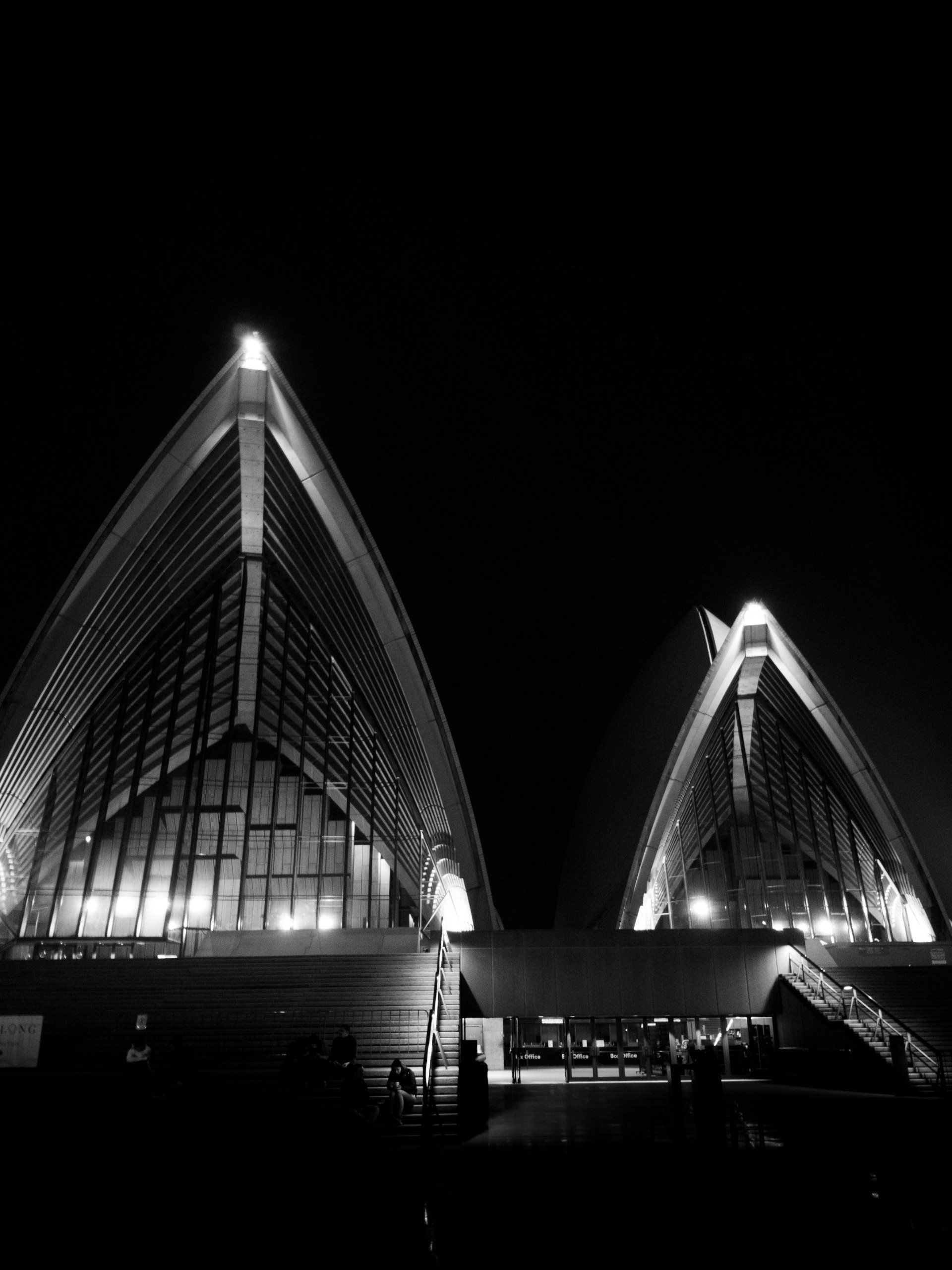 Ópera de Sydney à noite, em preto e branco. Velas arquitetônicas icônicas iluminadas contra um céu escuro.