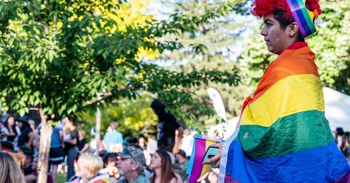 A man wearing a rainbow hat and a rainbow flag is standing in front of a crowd of people.