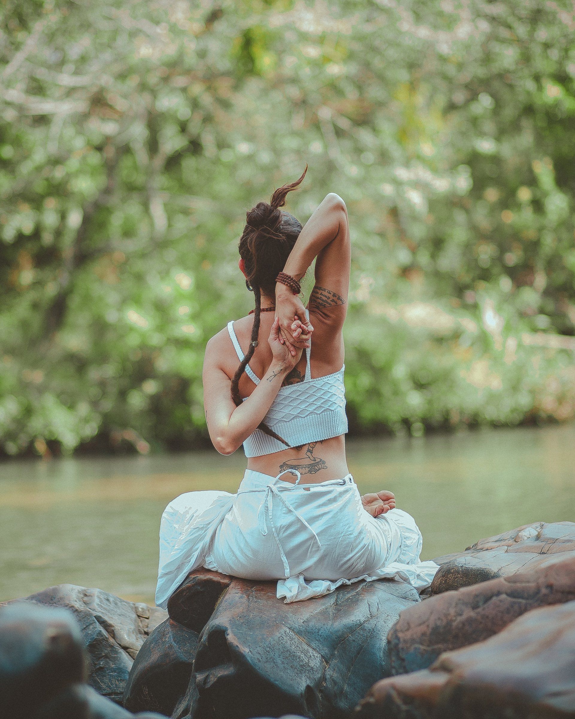 Woman in white yoga attire, arms behind back, seated on rock near water, outdoors.