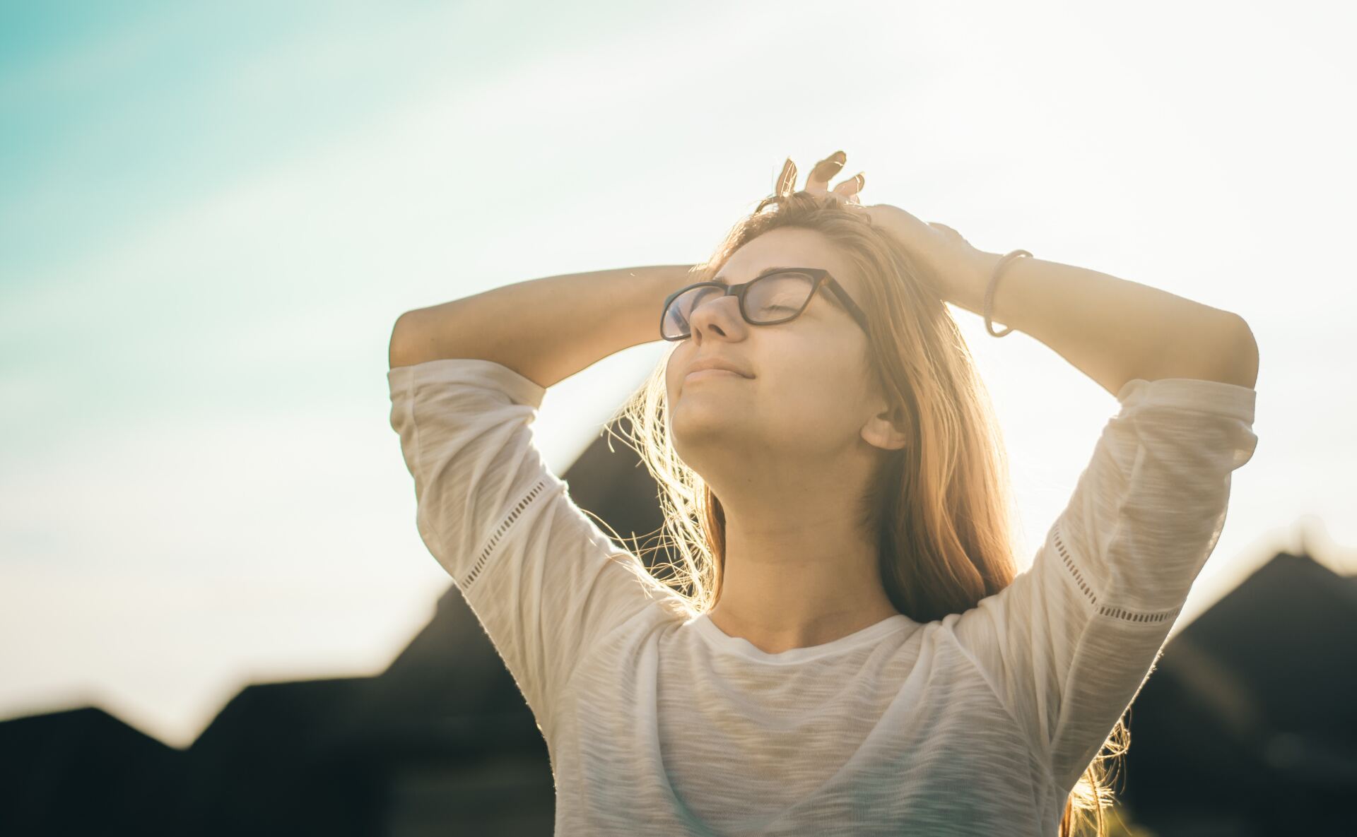 A woman wearing glasses is standing in the sun with her hands behind her head.