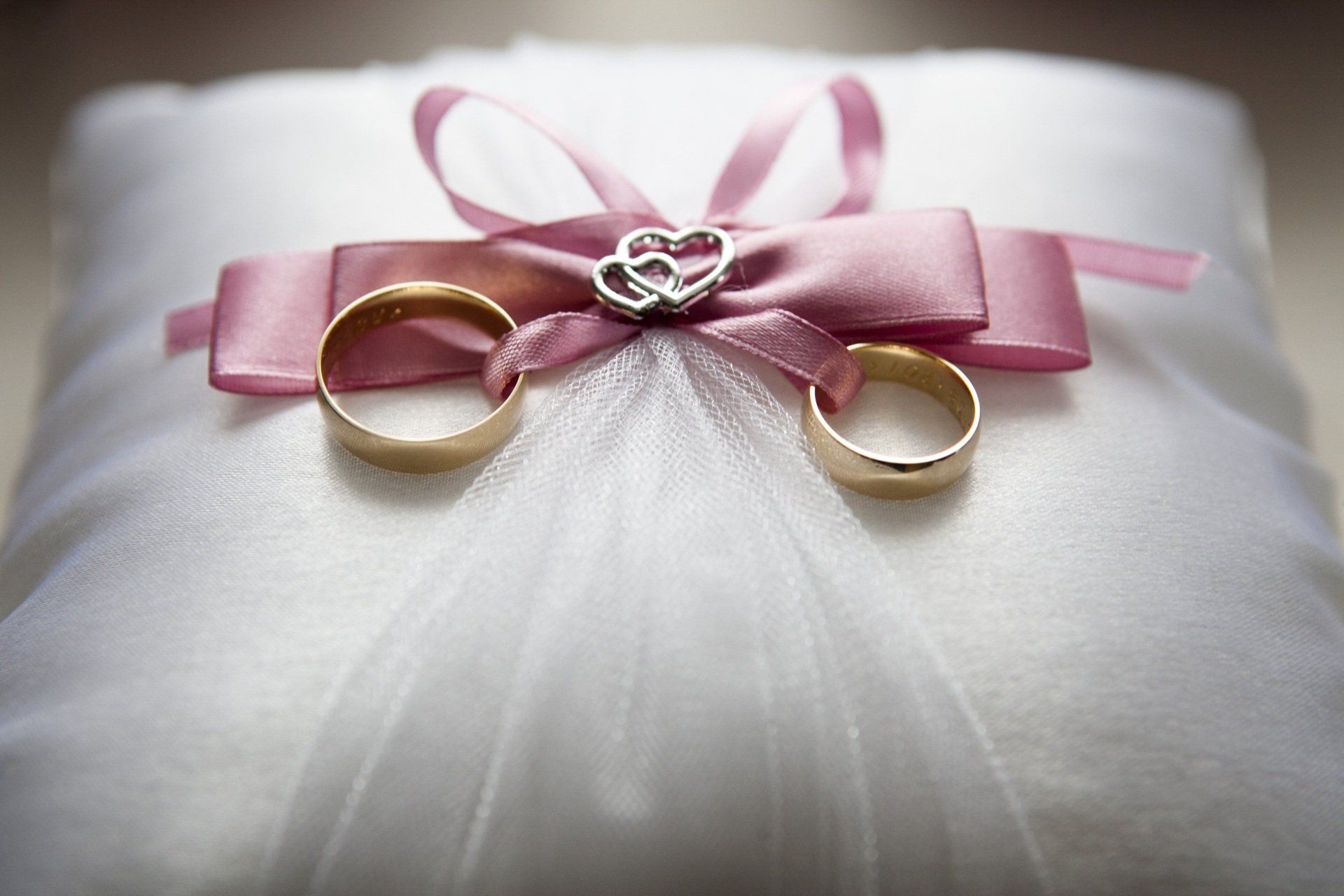 Two wedding rings are sitting on a white pillow with a pink bow.