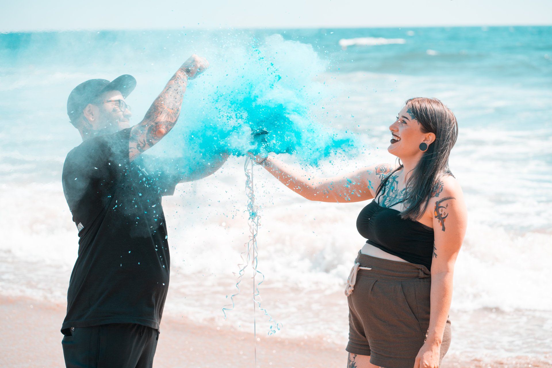 Pregnant couple on beach, reveal blue powder, happy expressions.