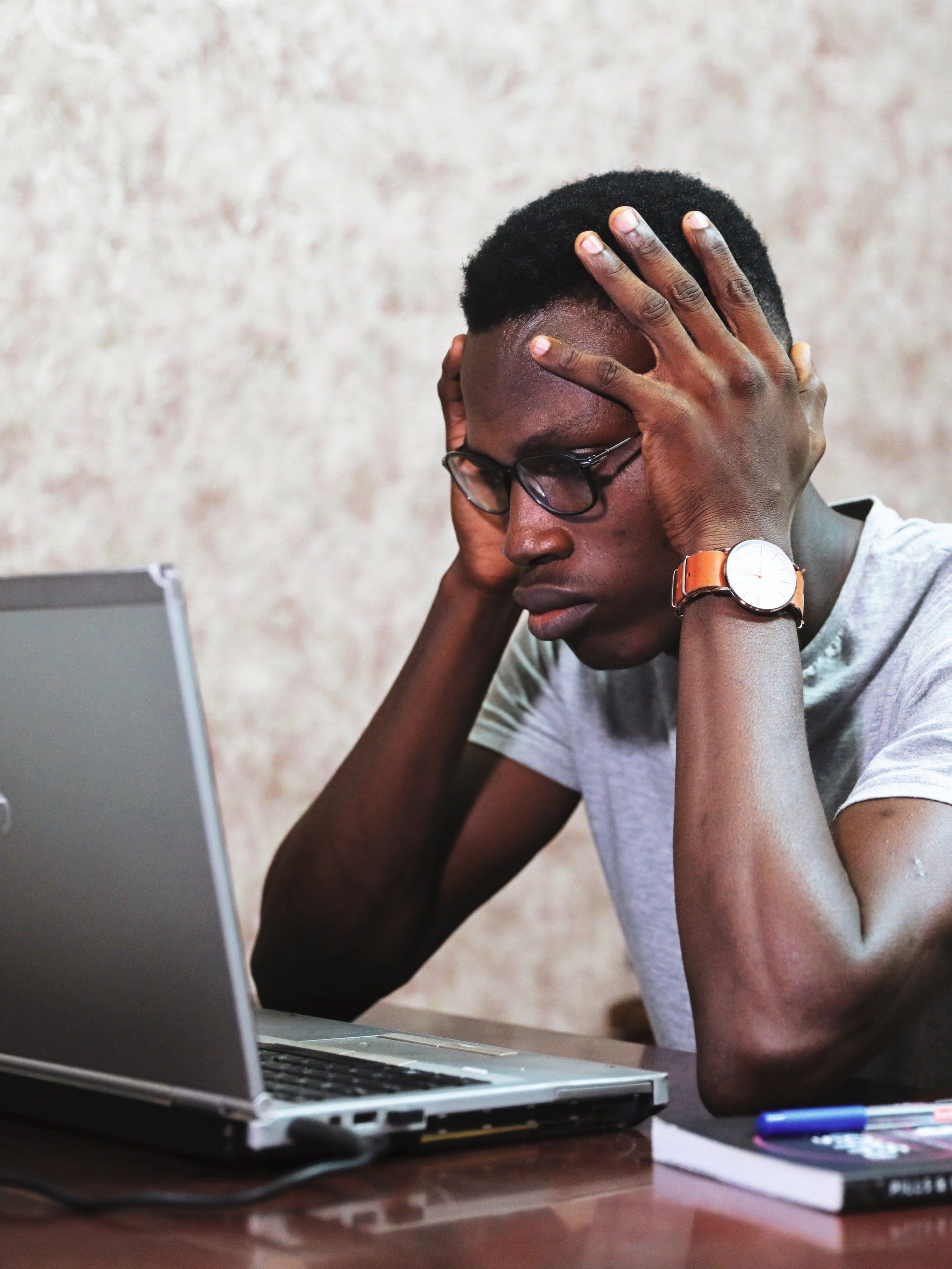 A man is sitting at a desk with his hands on his head looking at a laptop.