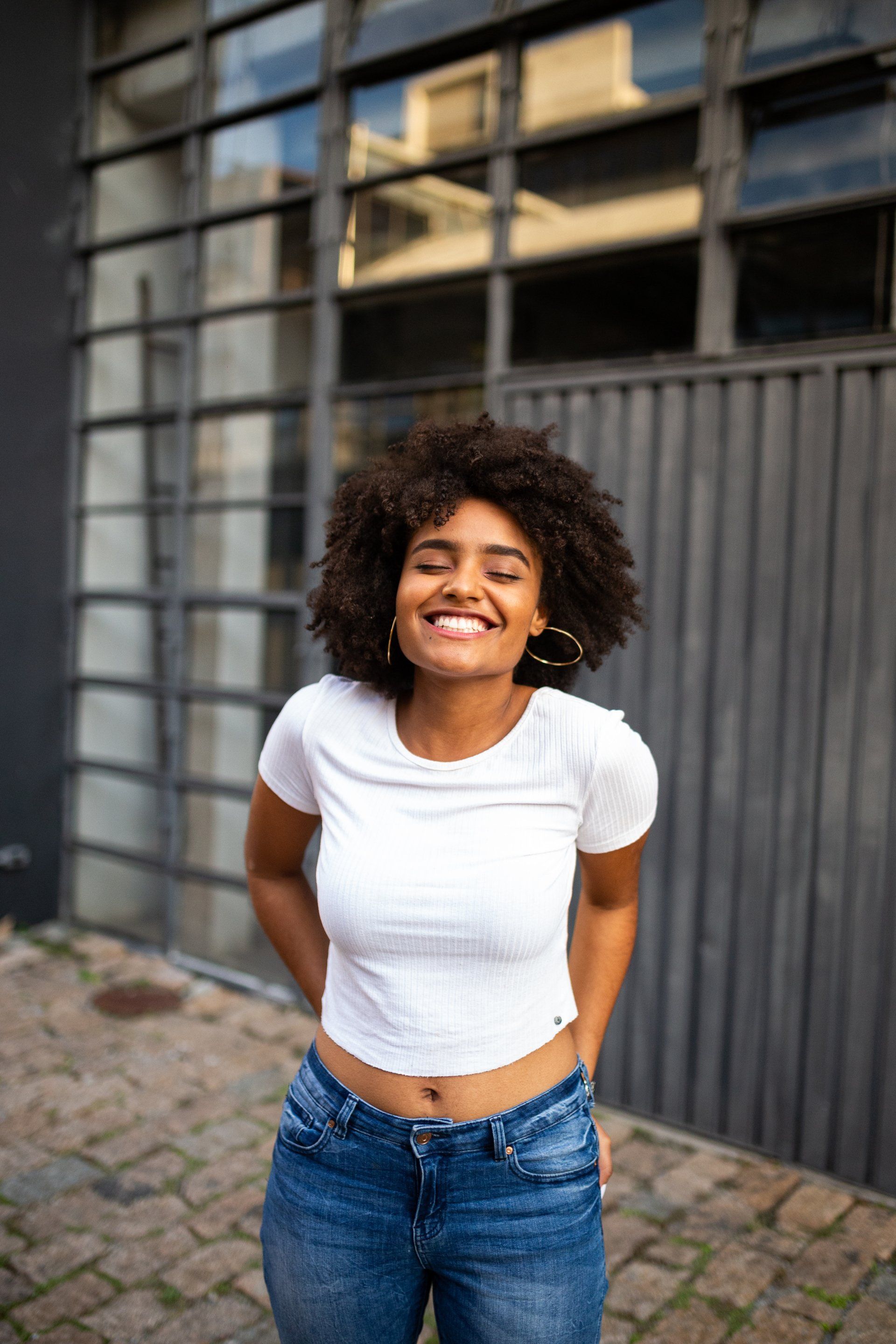 A woman in a white crop top and blue jeans is smiling in front of a building.