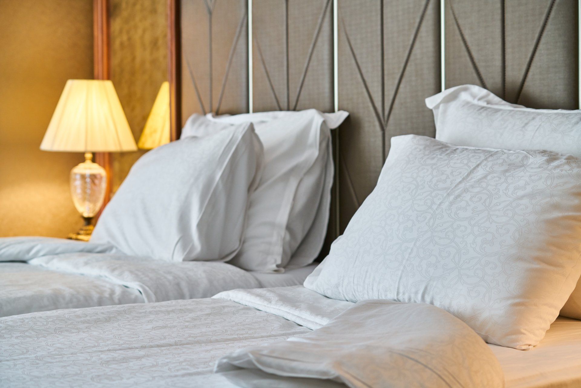 Duas camas brancas com roupa de cama branca, travesseiros e um abajur aceso sobre a mesa de cabeceira.