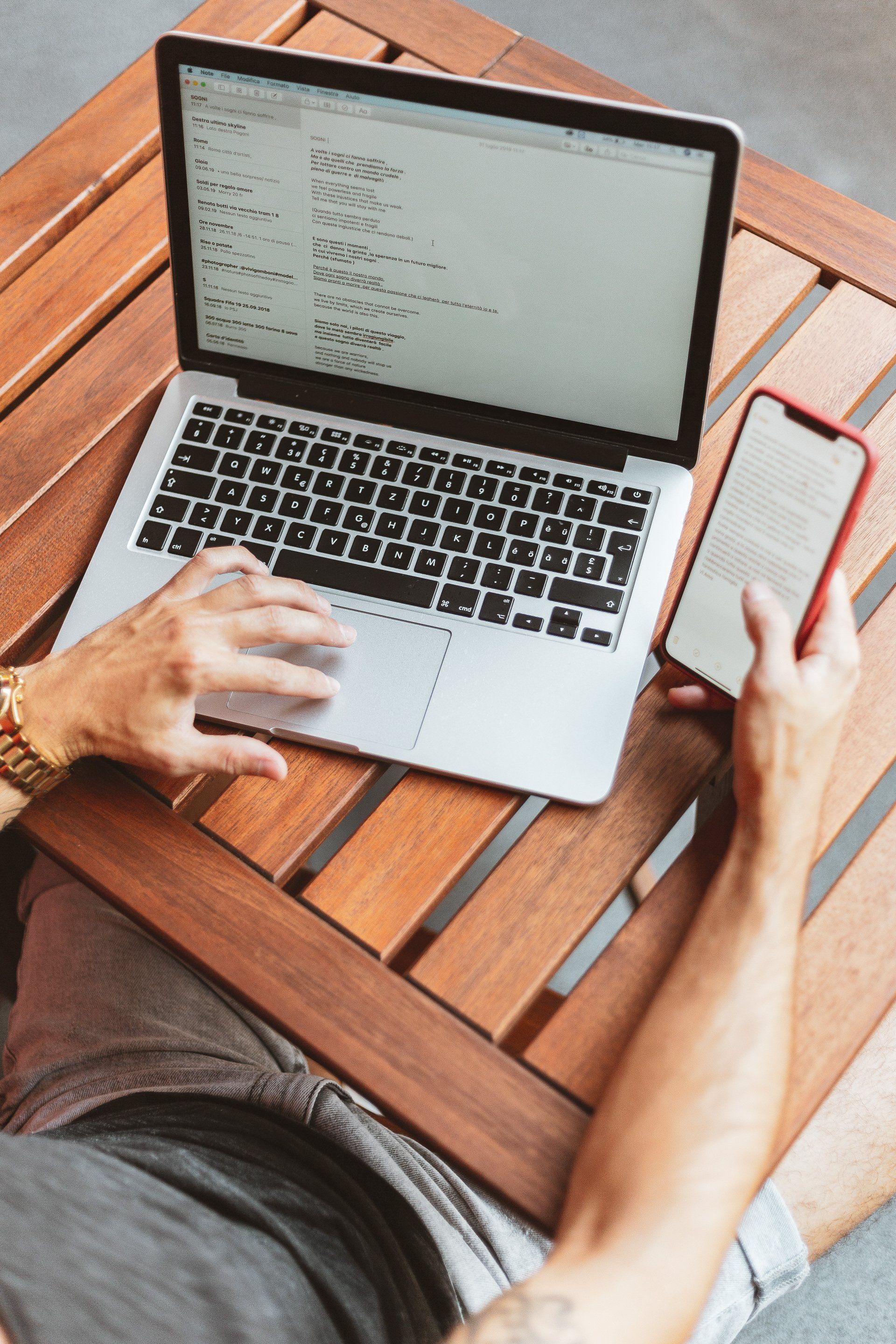 A man is sitting at a wooden table using a laptop and a cell phone.