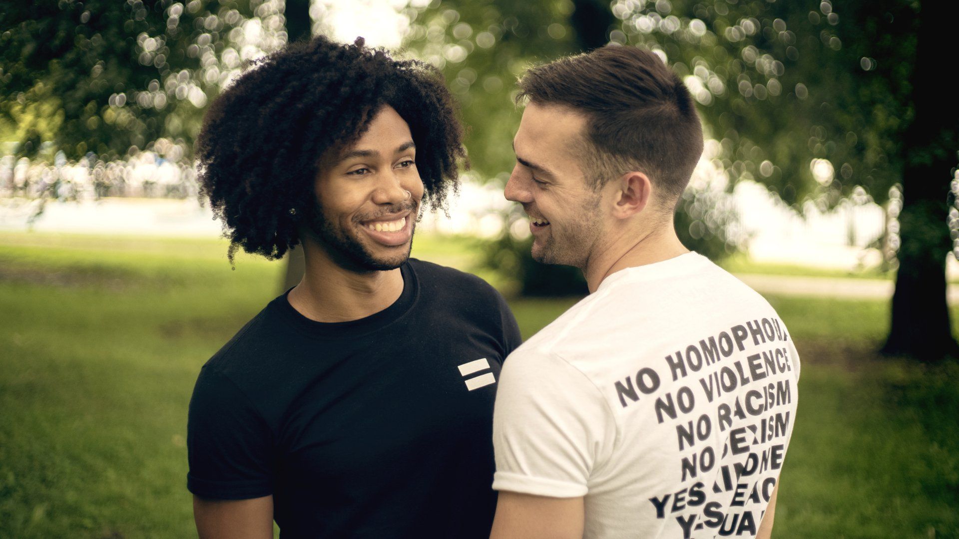 Two men are standing next to each other in a park and one of them is wearing a shirt that says no homophobia.