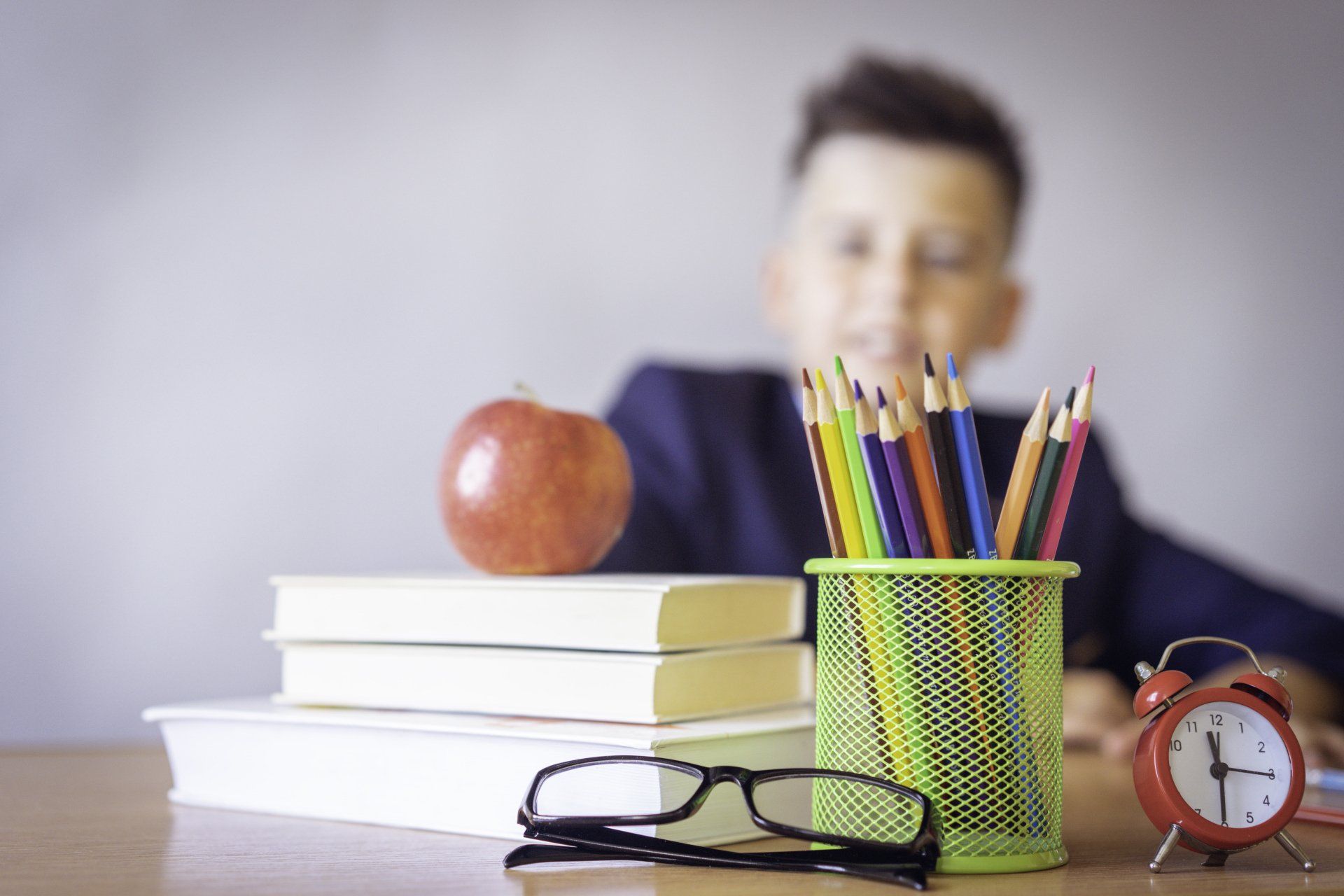 A boy is sitting at a desk with books , pencils , glasses and an apple.