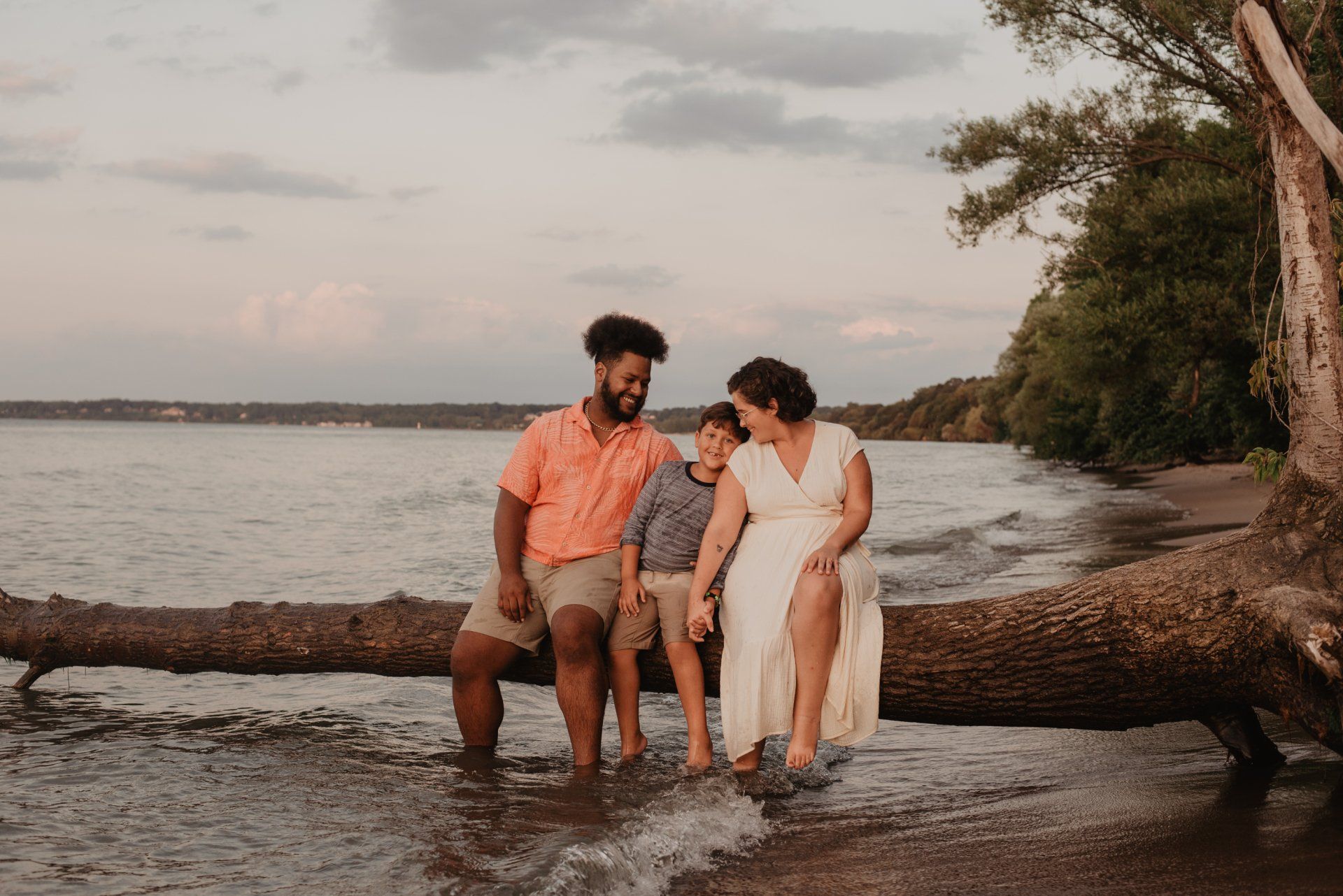 Family of three sitting on a fallen log at the water's edge. Golden light, waves, and trees.