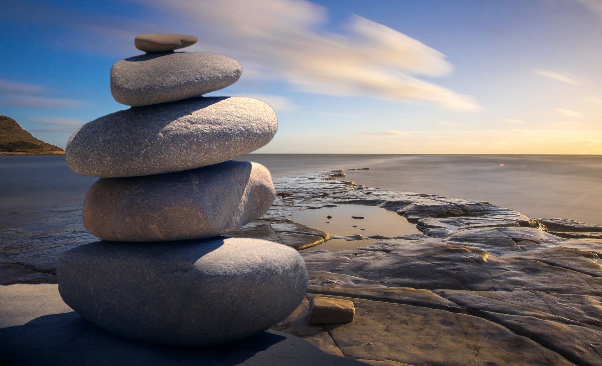 The image depicts a stacked rock Cairn, with five rocks arranged from largest at the bottom to smallest on the top, positioned on the edge of an ocean view. This image represents my virtual wellness program called SoulQuest online. SoulQuest offers various levels of spiritual guidance focused on three key aspects: purpose, mindfulness, and connection. The program aims to collaborate with clients in envisioning their greater life purpose and providing them with practical tools and methods to cultivate awareness, enlightenment, and meaningful experiences.