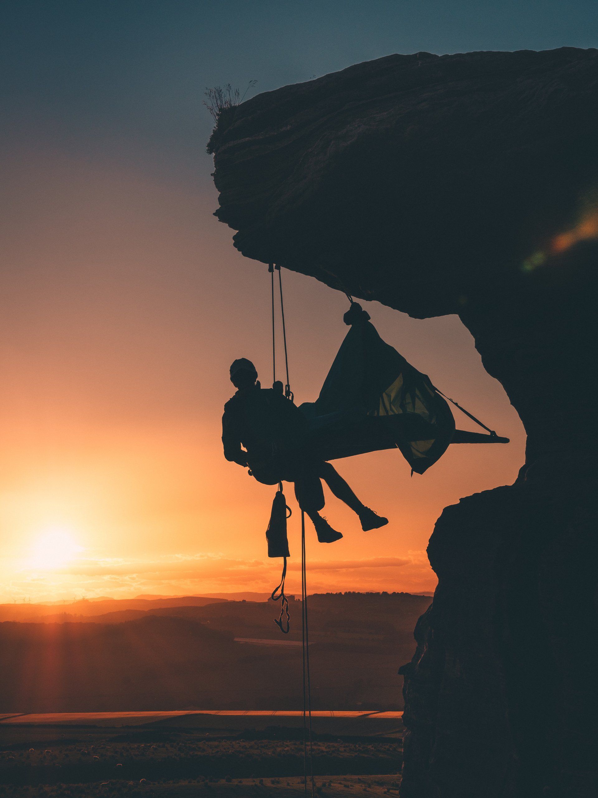 a person is sitting in a tent on a cliff at sunset with a rescue worker using ropes t access.