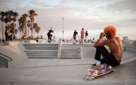 A skateboarder films as his friend ollies a stair set at a skatepark.