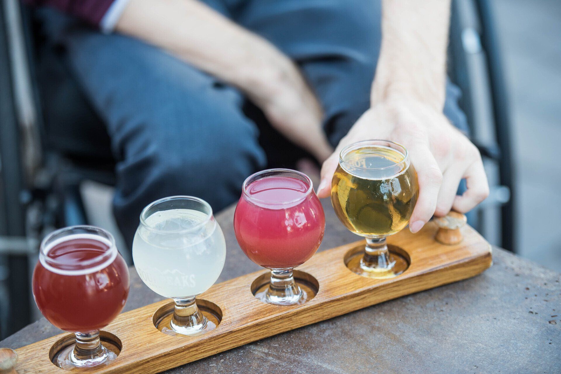 A person in a wheelchair is holding a wooden tray with four glasses of beer on it.