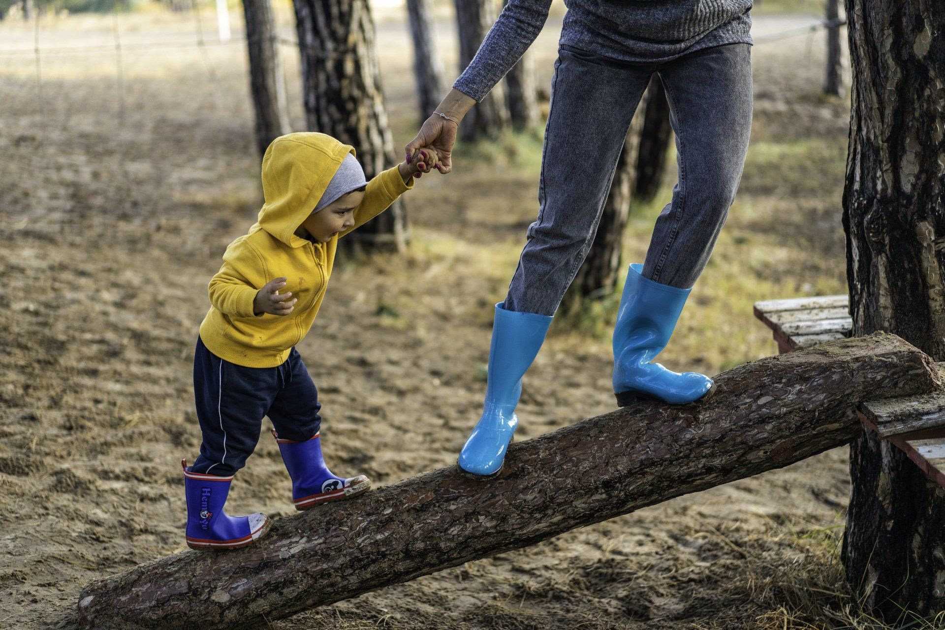 A woman is helping a little boy climb a log in the woods.