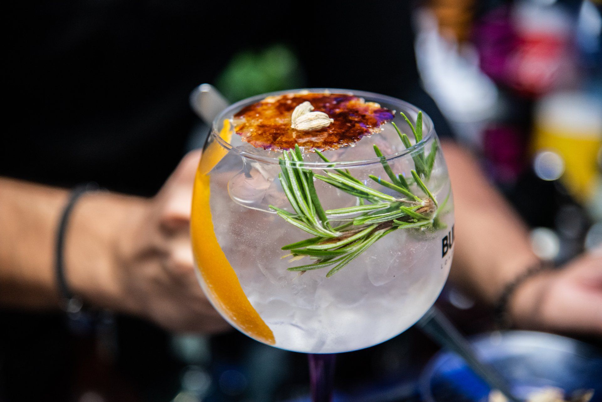 A cocktail in a clear glass with rosemary and an orange peel garnish, being made by a bartender.