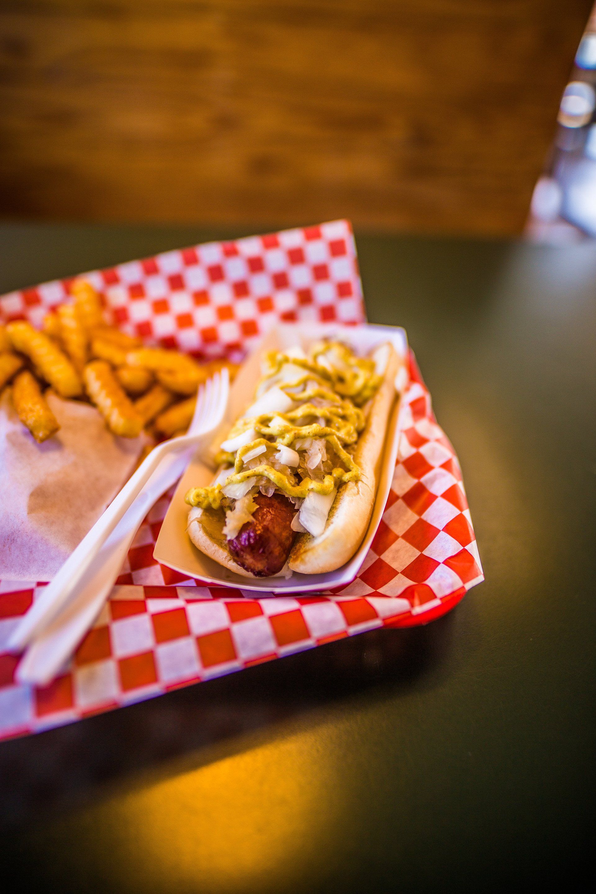 Hot dog with mustard and onions in a tray with fries and a fork. Red and white checkered paper.