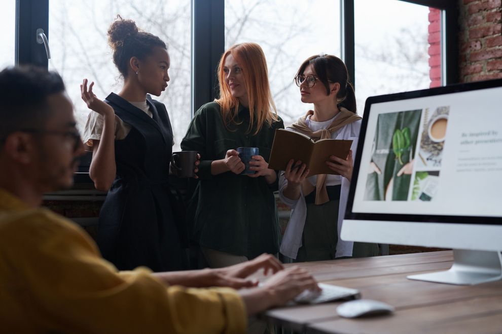 image of three women drinking coffee in front of another woman on a computer