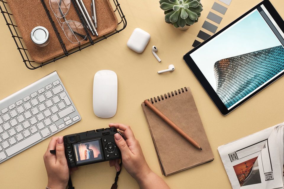 Hands holding camera over desk with keyboard, mouse, tablet, notebook, and accessories.