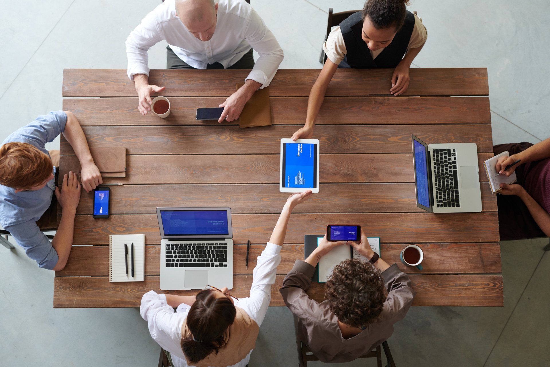 A group of people are sitting around a wooden table with laptops and tablets.