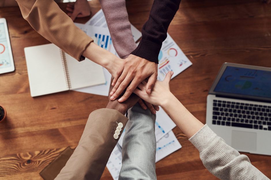 Hands piled together in a team huddle over a wooden table with documents and a laptop, signifying teamwork and collaboration.