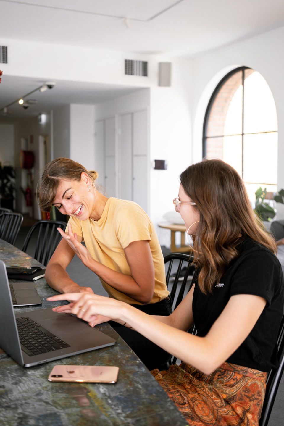 Two women laughing while looking at a laptop in a well-lit office. One points, the other gestures.