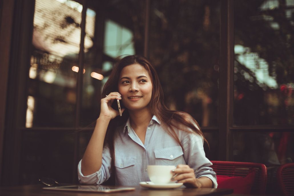 A woman is sitting at a table talking on a cell phone while holding a cup of coffee.