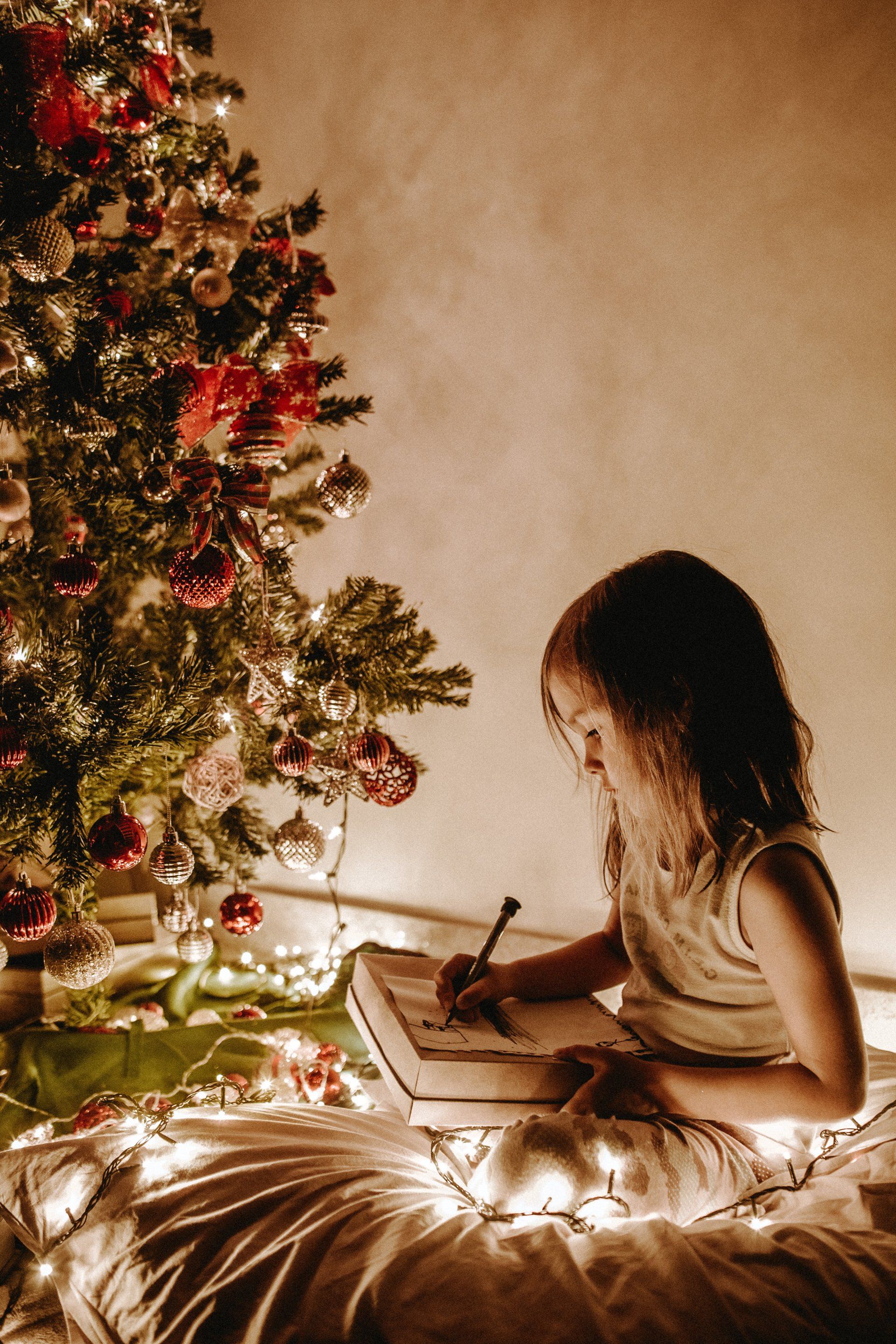 A little girl is sitting in front of a christmas tree writing in a notebook.