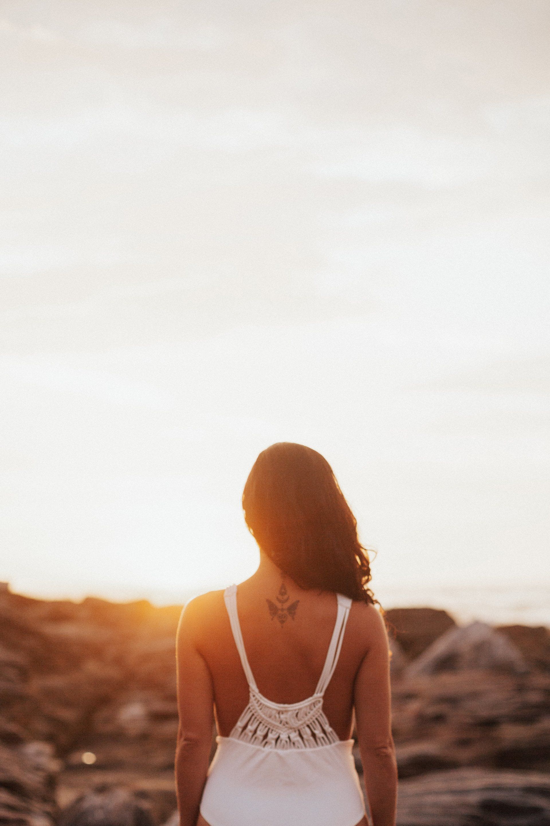 A woman in a white swimsuit is standing on a rocky beach at sunset.