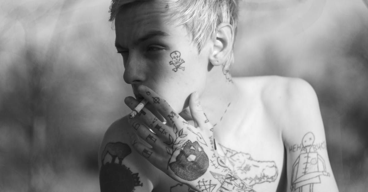Homme avec des tatouages sur le visage et les mains, fumant une cigarette ; noir et blanc.
