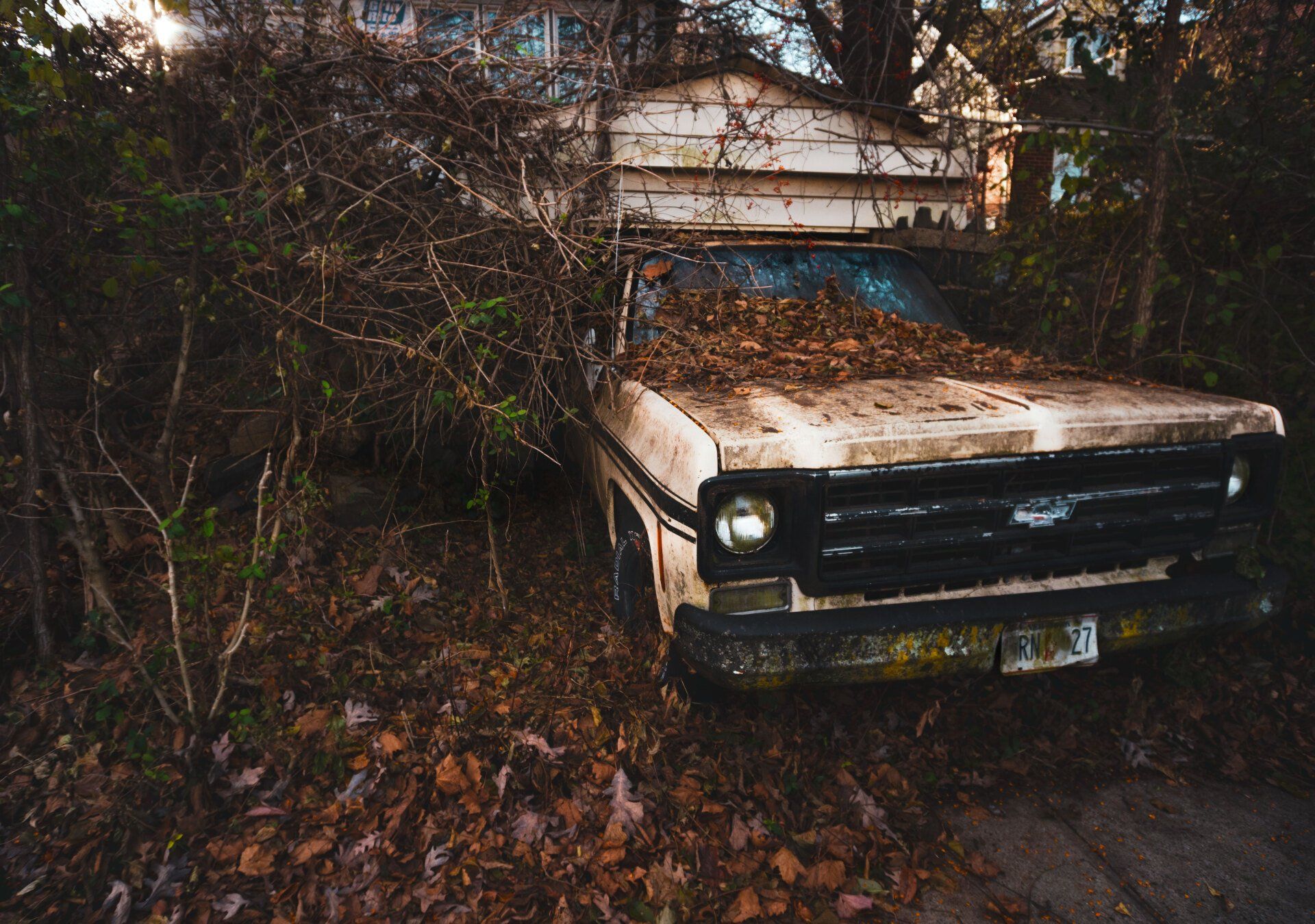 An old truck is covered in leaves in front of a house.