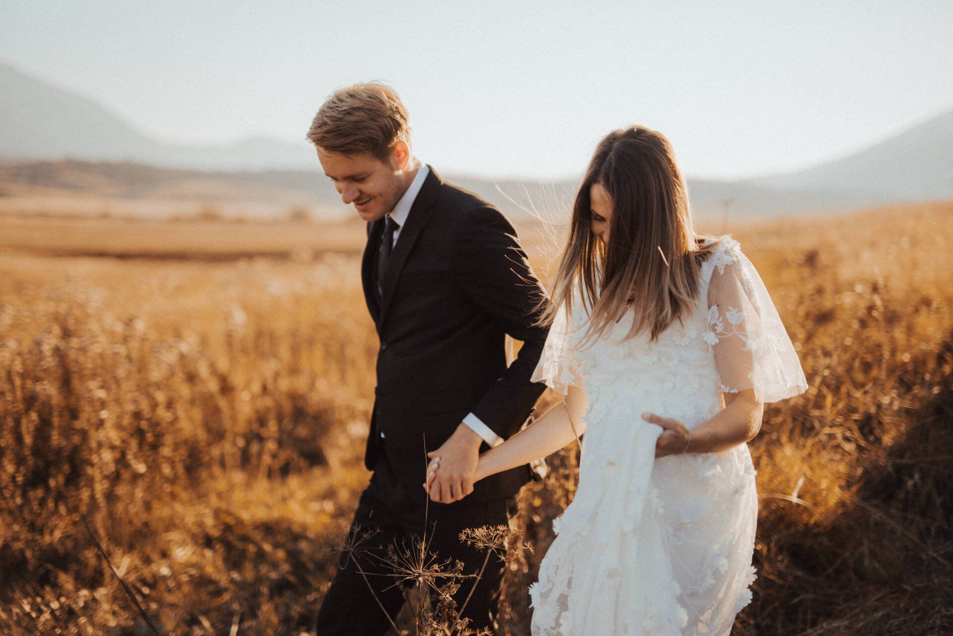 a bride and groom are walking through a field holding hands 