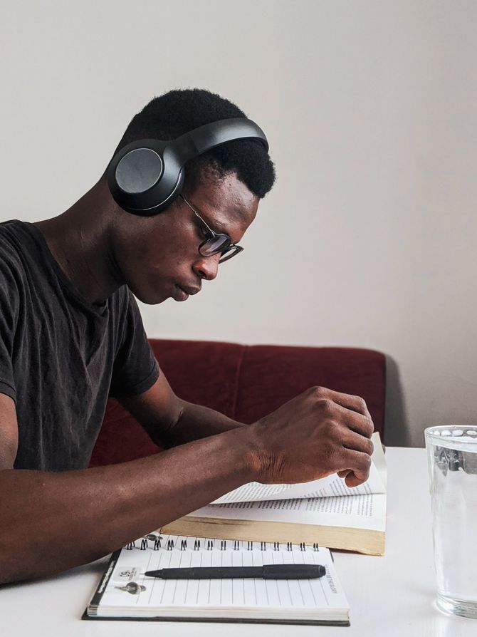 A man wearing headphones is sitting at a table writing in a notebook.
